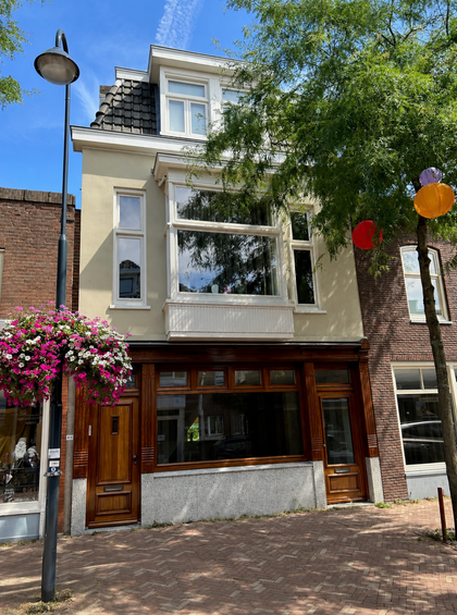 Smalle bakstenen gevel van een rijtjeshuis met grote ramen, houten deur en hangende bloemen aan een zonnige straat.