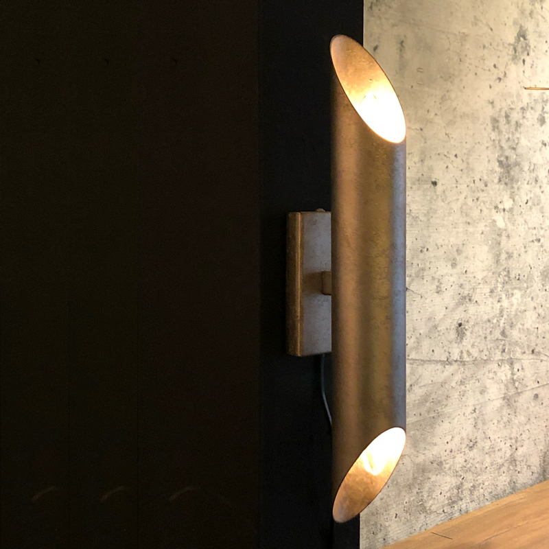 Een wandlamp met twee lampjes, bronskleurig, aan een donkere muur met een betonachtige uitstraling.