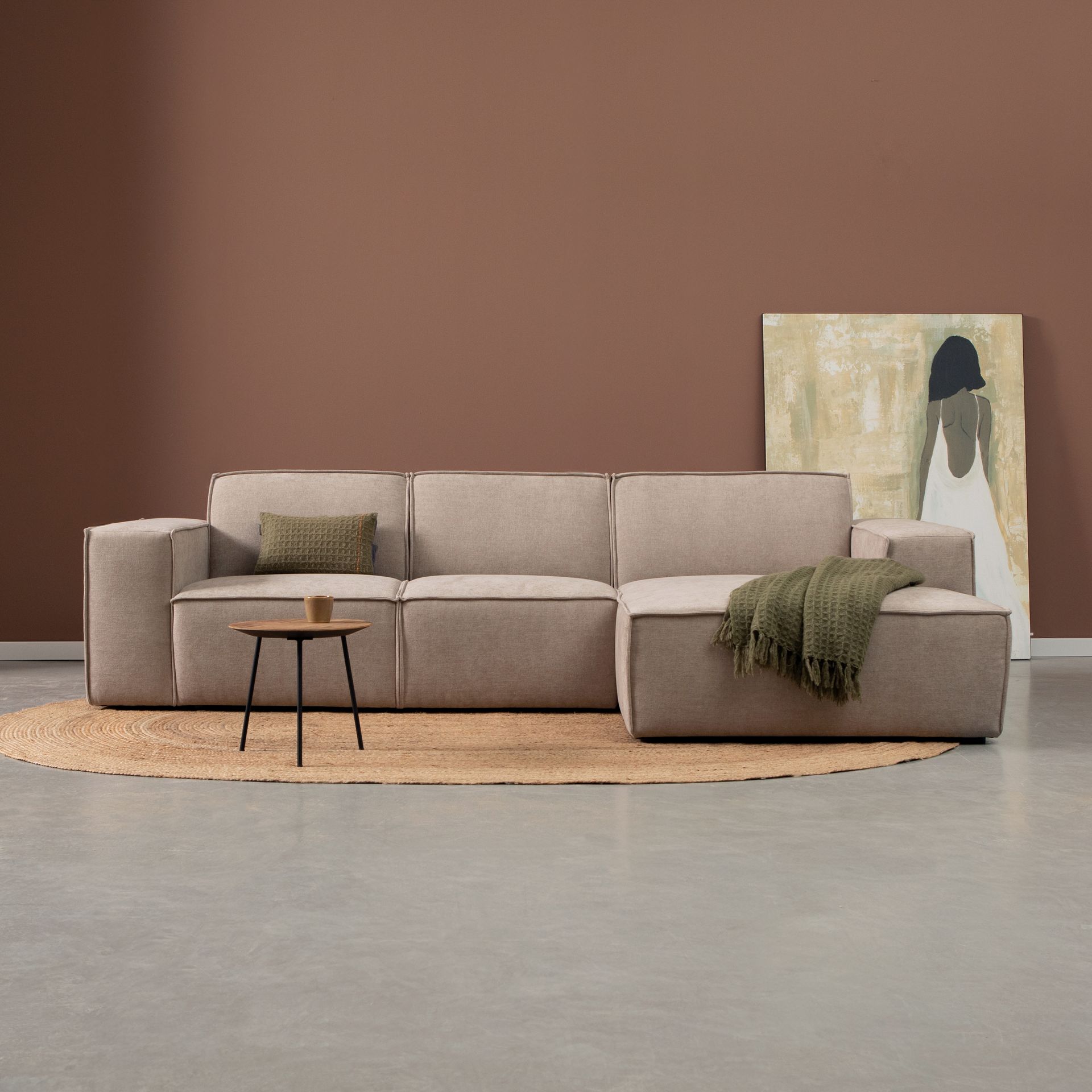 Moderne woonkamer met beige hoekbank, ronde salontafel, geweven vloerkleed en abstracte kunst tegen een bruine muur.