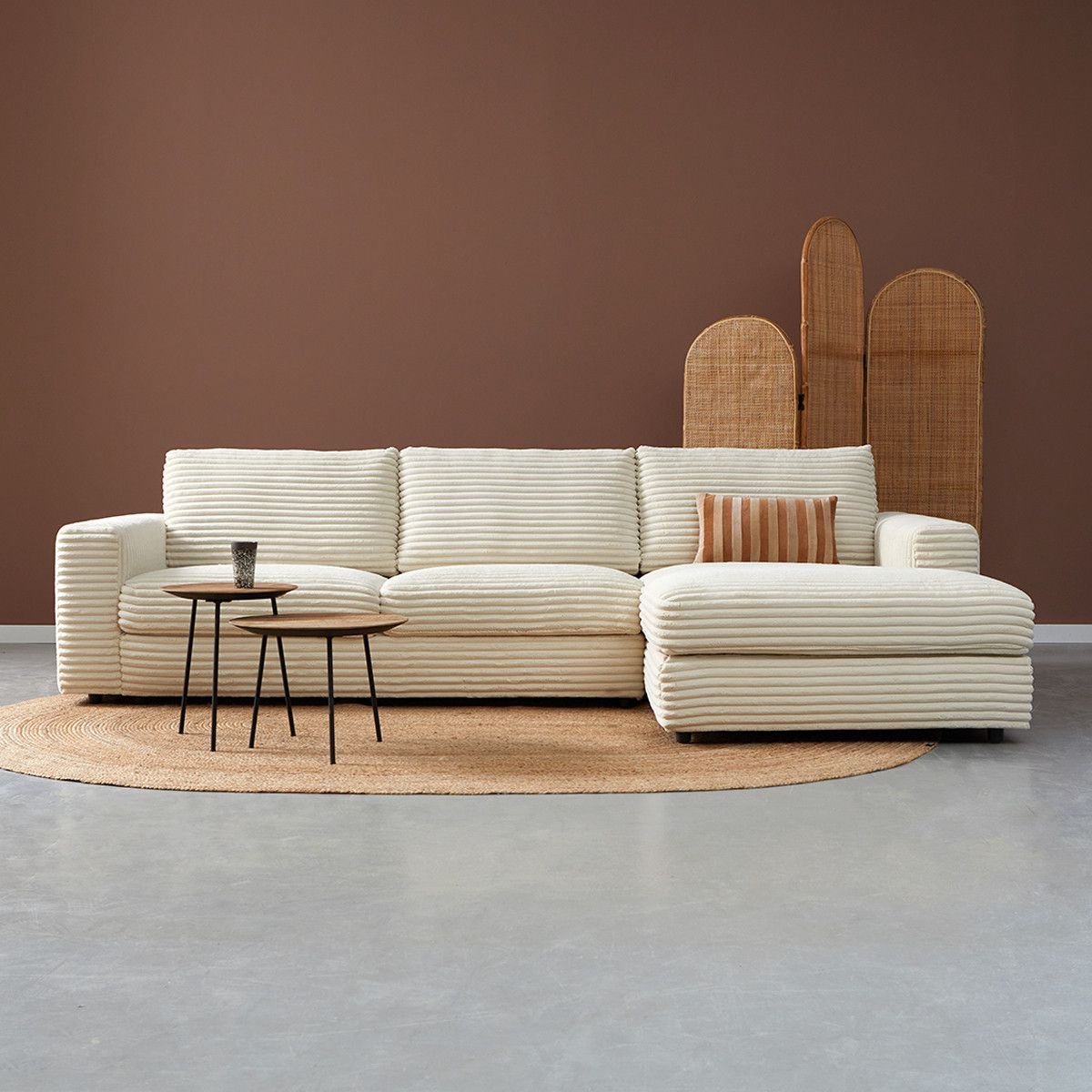 Moderne beige gestreepte hoekbank met salontafels op een rond vloerkleed tegen een bruine muur.