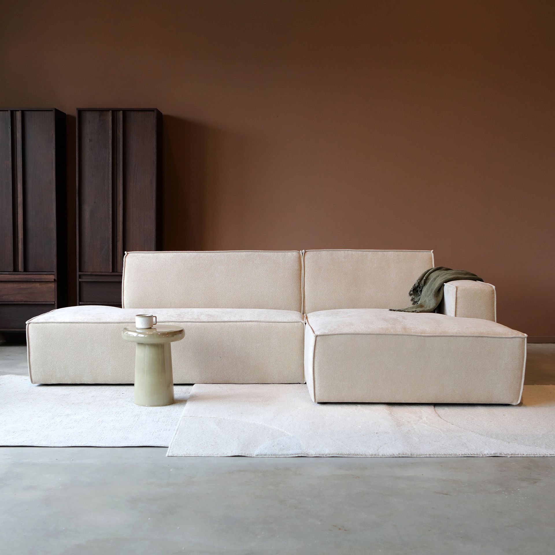 Een minimalistische beige hoekbank in een moderne kamer met een klein rond bijzettafeltje en donkere kasten.