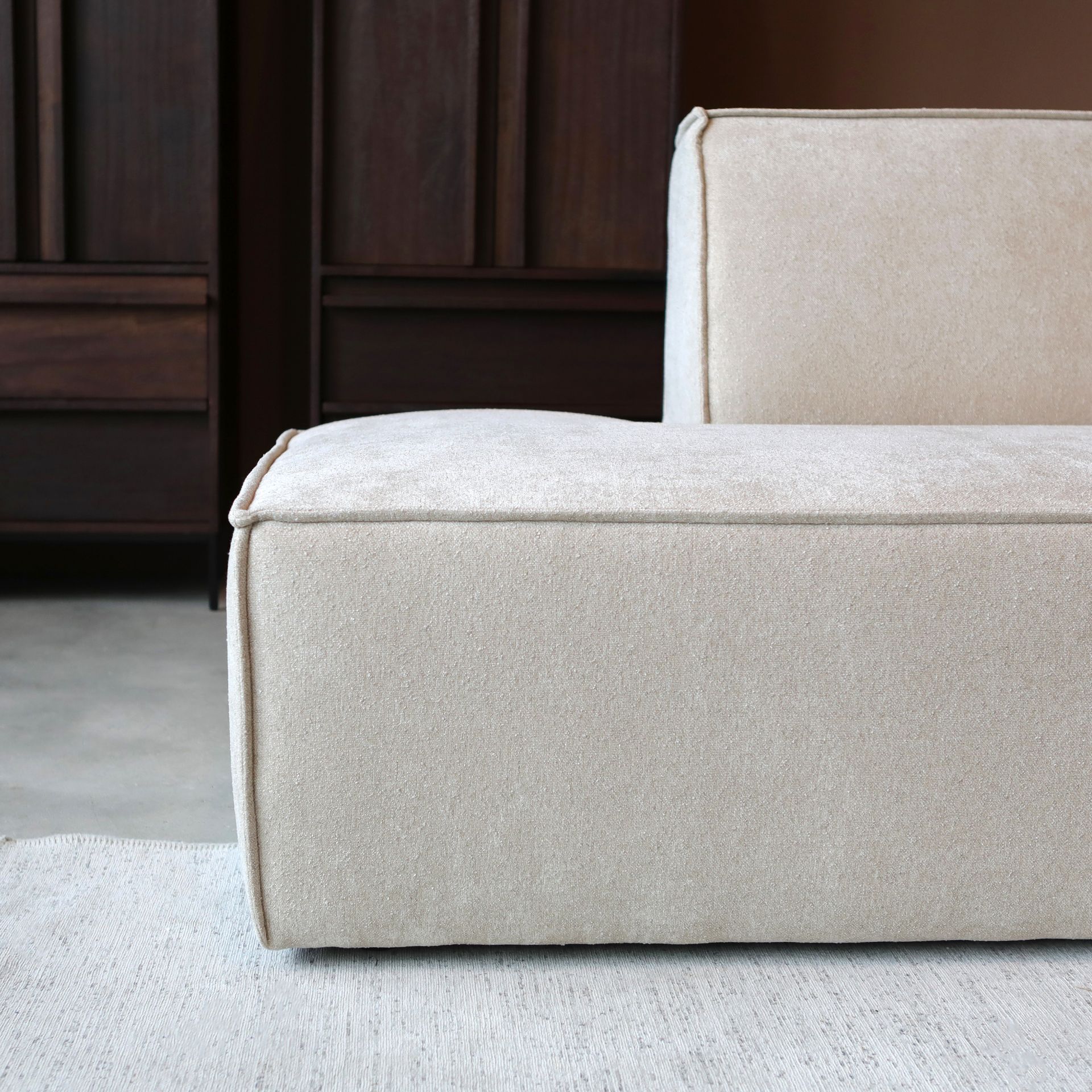 Een minimalistische beige hoekbank in een moderne kamer met een klein rond bijzettafeltje en donkere kasten.