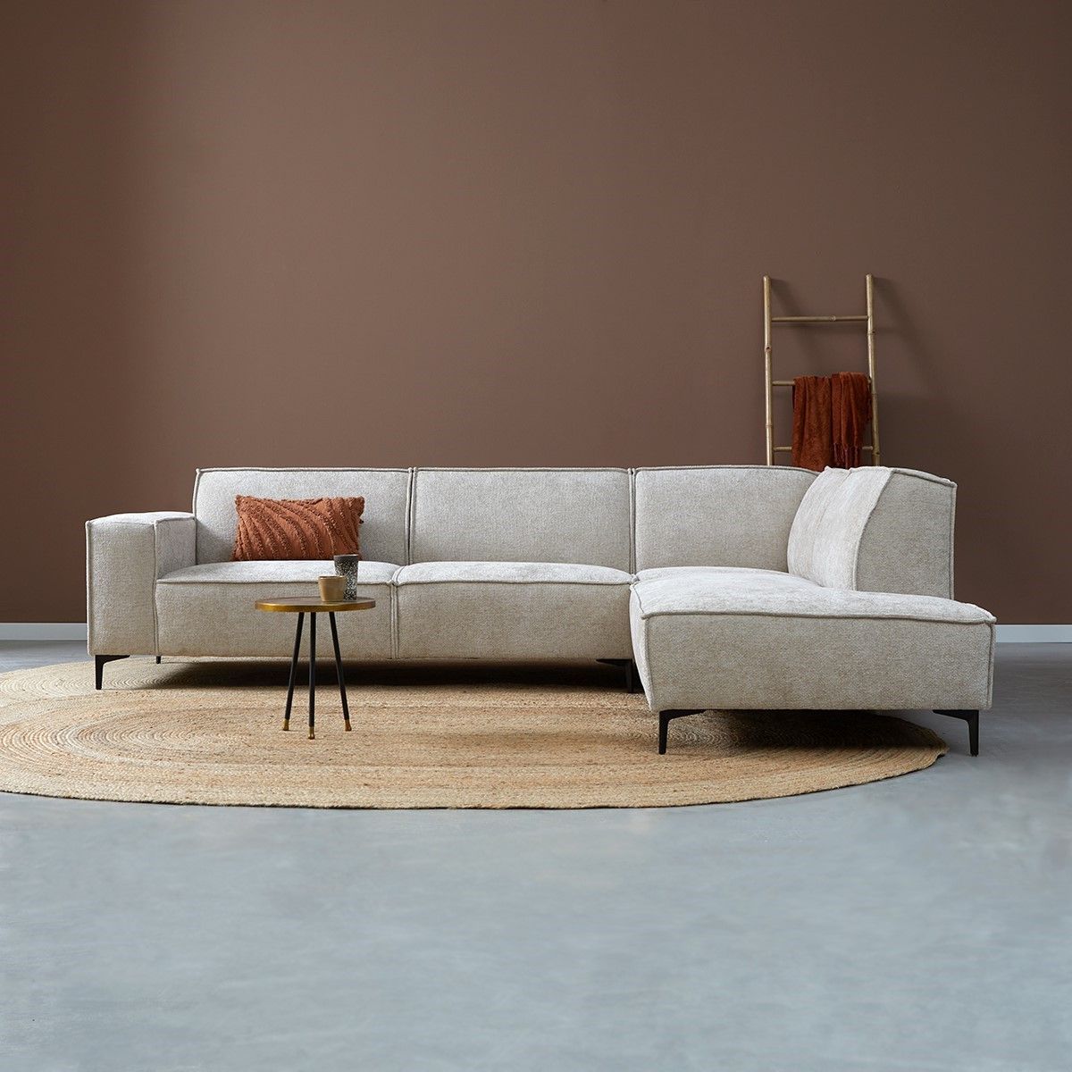 Moderne woonkamer met een lichtgrijze hoekbank, een kleine ronde tafel en bruine wanddecoratie op een beige vloerkleed.