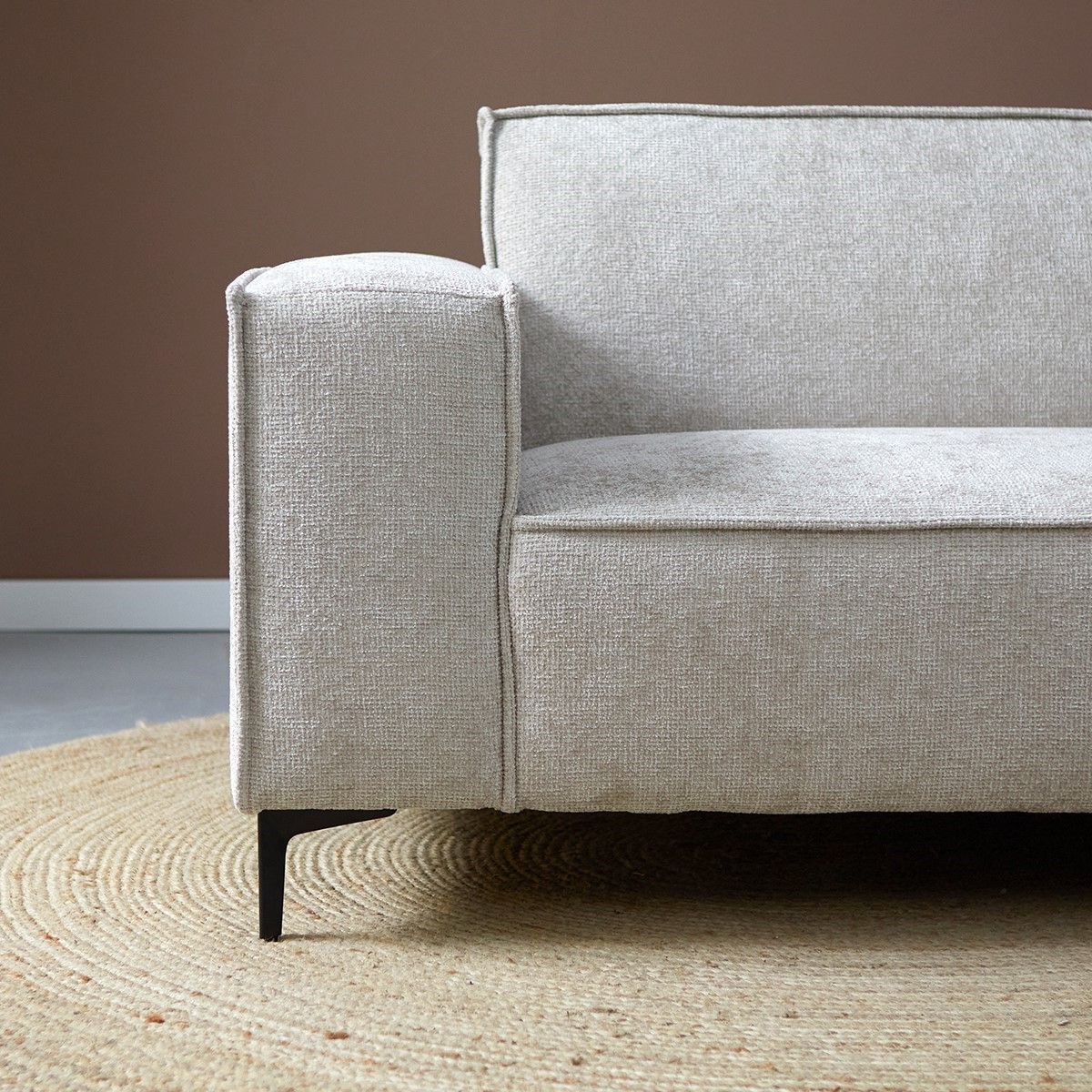 Moderne woonkamer met een lichtgrijze hoekbank, een kleine ronde tafel en bruine wanddecoratie op een beige vloerkleed.