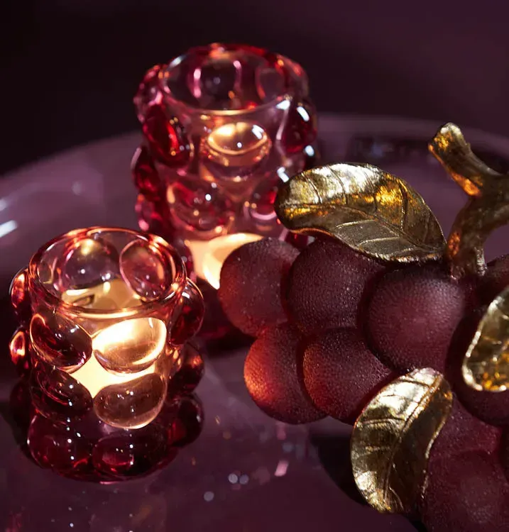 Twee roze glazen kaarsenhouders en een decoratieve druiventros op een paarse ondergrond, verlicht door warm licht.