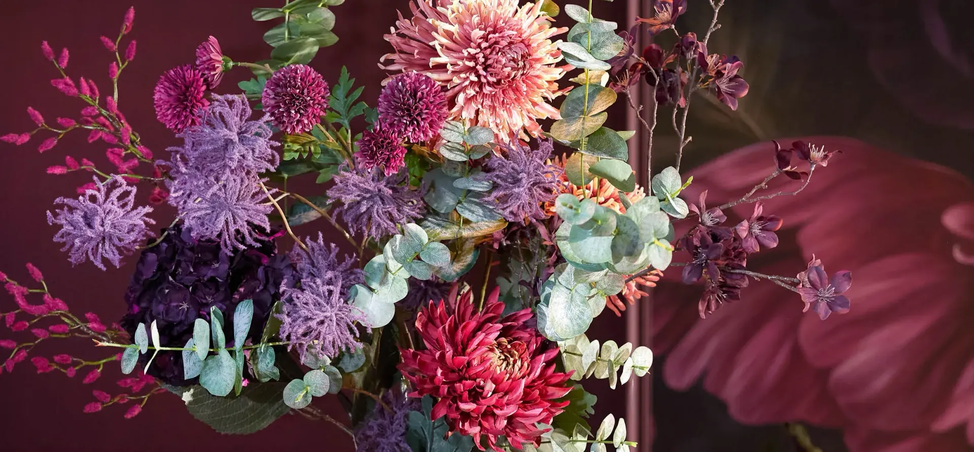 Een levendig bloemstuk met diverse gekleurde bloemen en groen tegen een dieprode achtergrond.