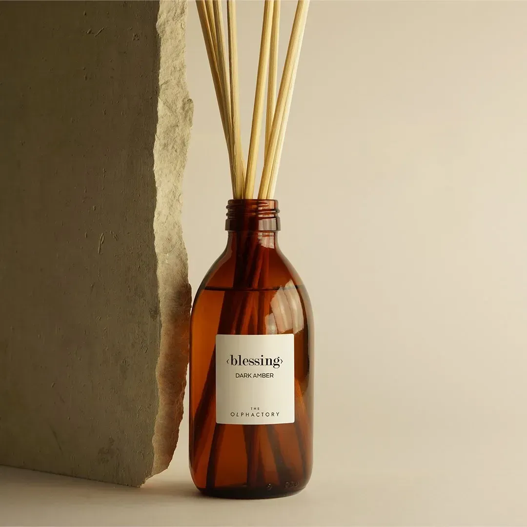 Amberkleurige rietdiffuserfles met beige rietjes naast een muur met structuur.