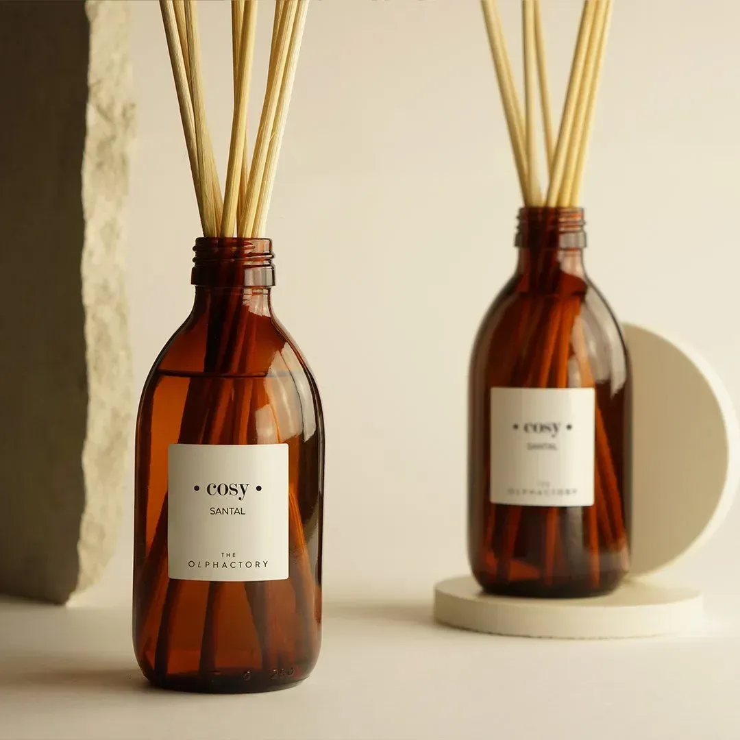 Amberkleurige rietdiffuserfles met gedroogde rietjes op een beige ondergrond tegen een muur met structuur.