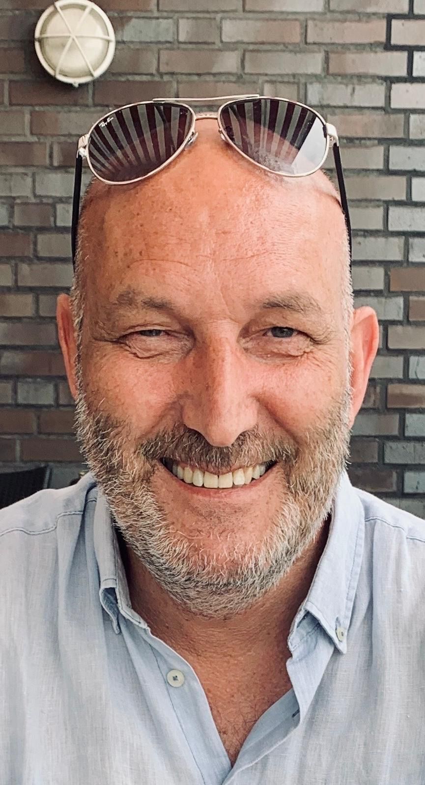 Een lachende man met een zonnebril op zijn hoofd staat voor een bakstenen muur.