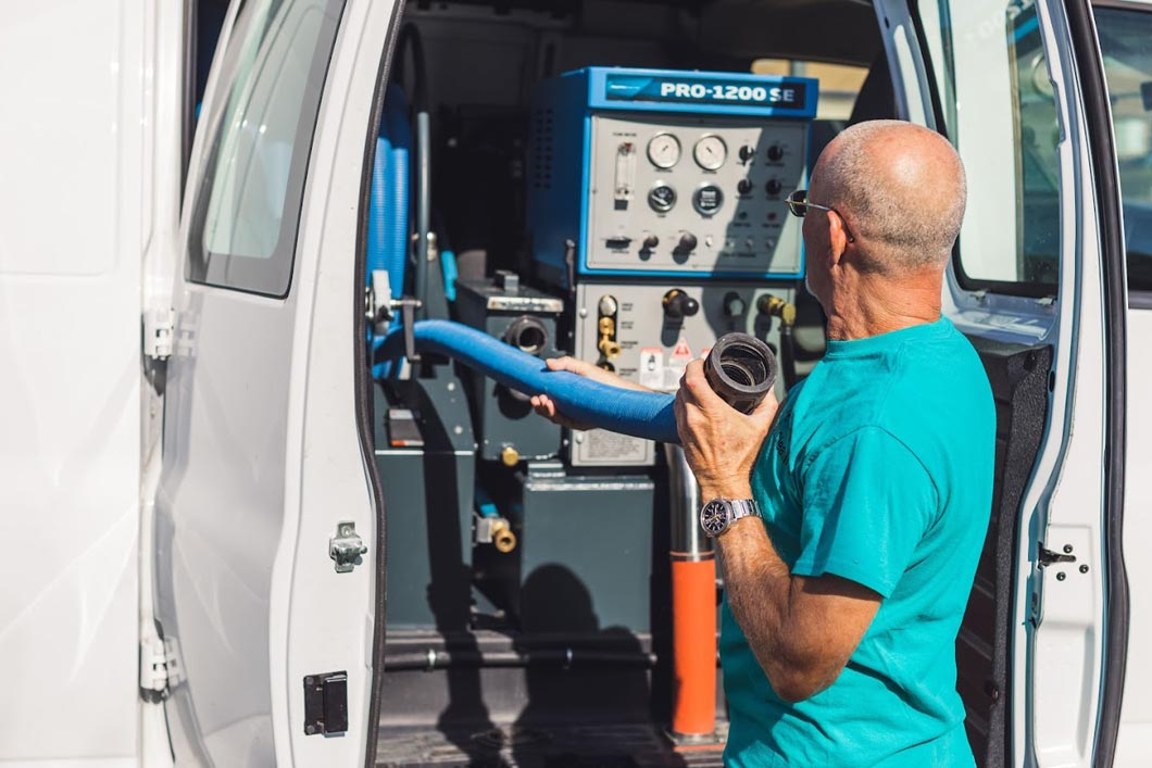 Man in a turquoise shirt near a van, holding a black nozzle, next to a blue cleaning machine.