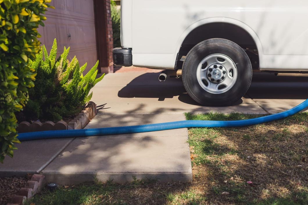 Blue hose extends from white van parked on a driveway with greenery.