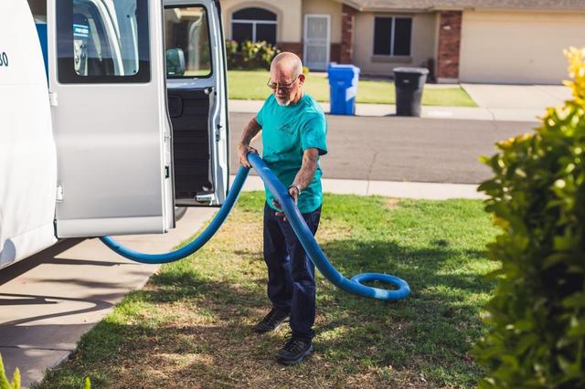 Man in teal shirt and dark pants, handling a blue hose, near a white van on a green lawn.