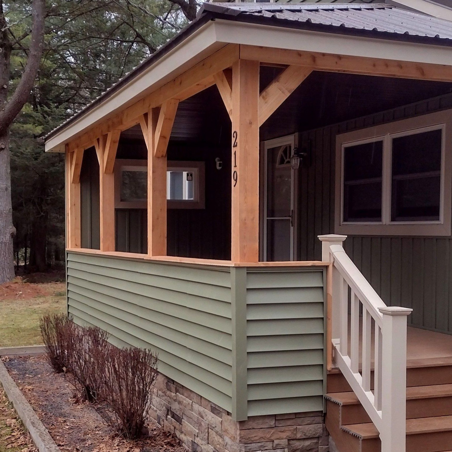 Covered porch with green siding, wood posts, white railing, and stone foundation on a house