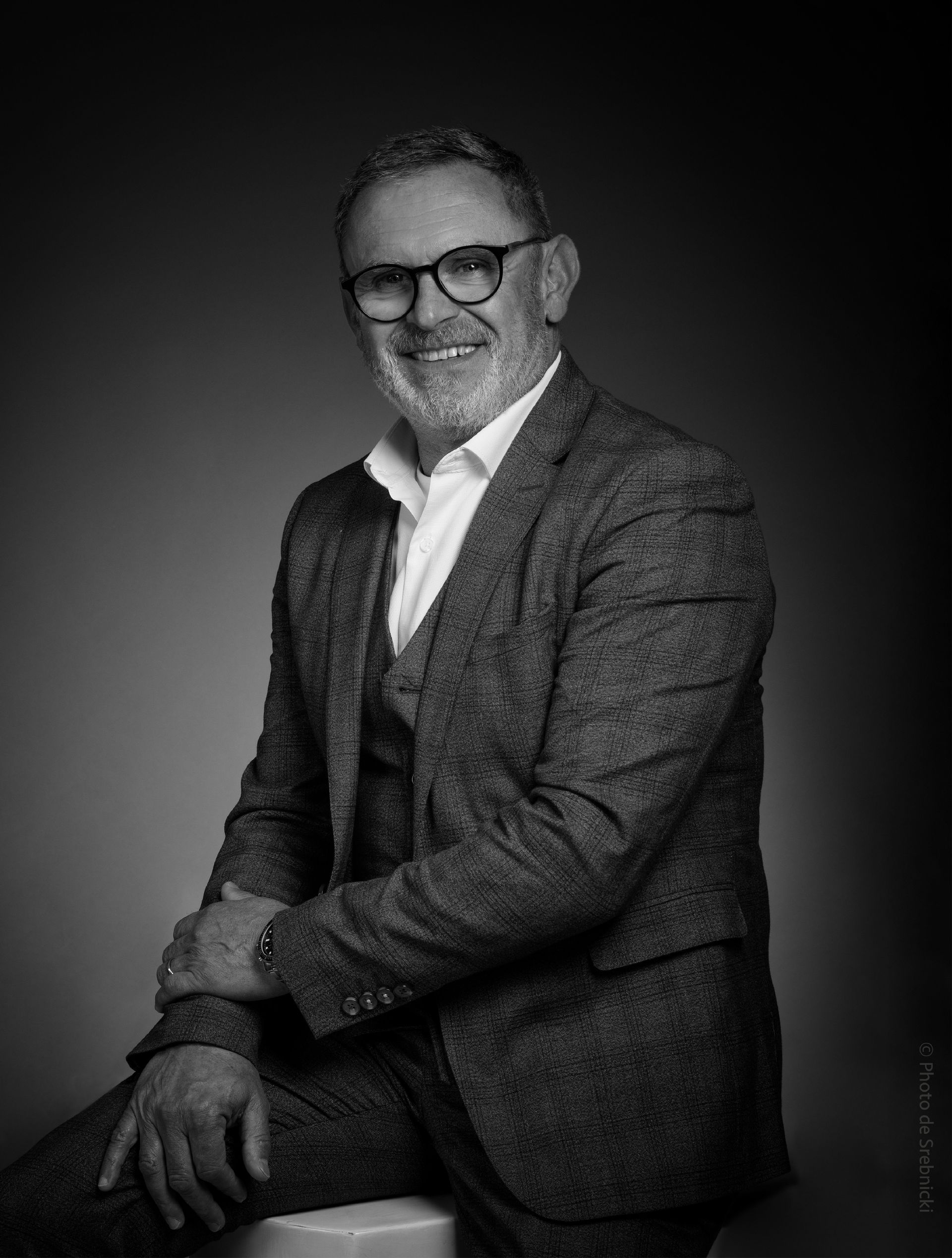 Un homme en costume et lunettes est assis sur un tabouret sur une photo en noir et blanc.
