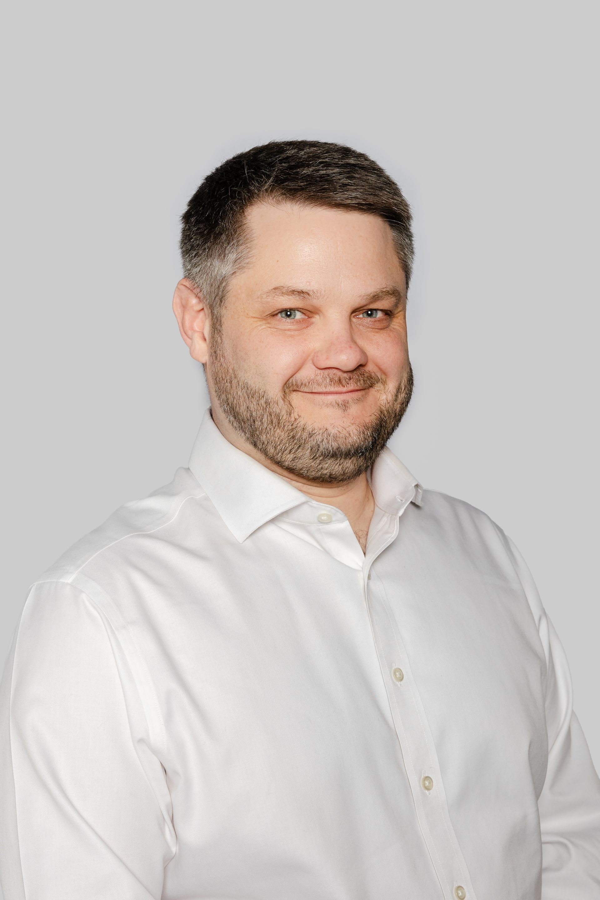 Une personne aux cheveux bruns courts et à la barbe, portant une chemise blanche boutonnée, souriant sur un fond gris clair.
