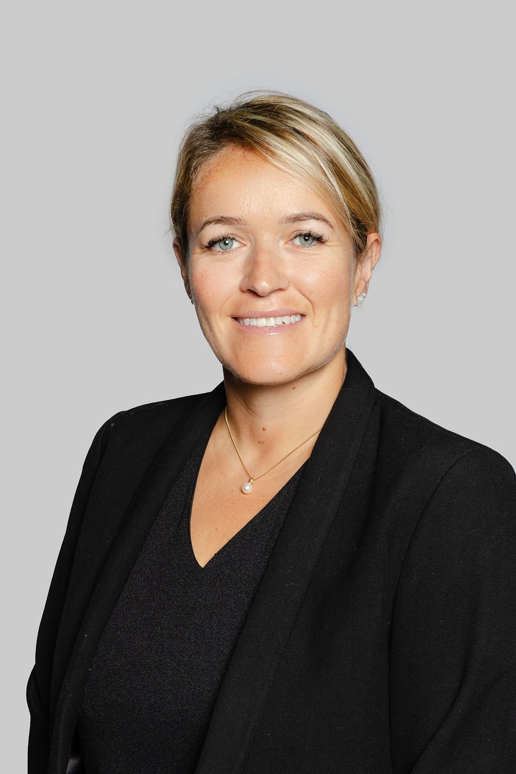 Femme aux cheveux blonds, souriante, portant un blazer noir et un collier, sur fond gris.