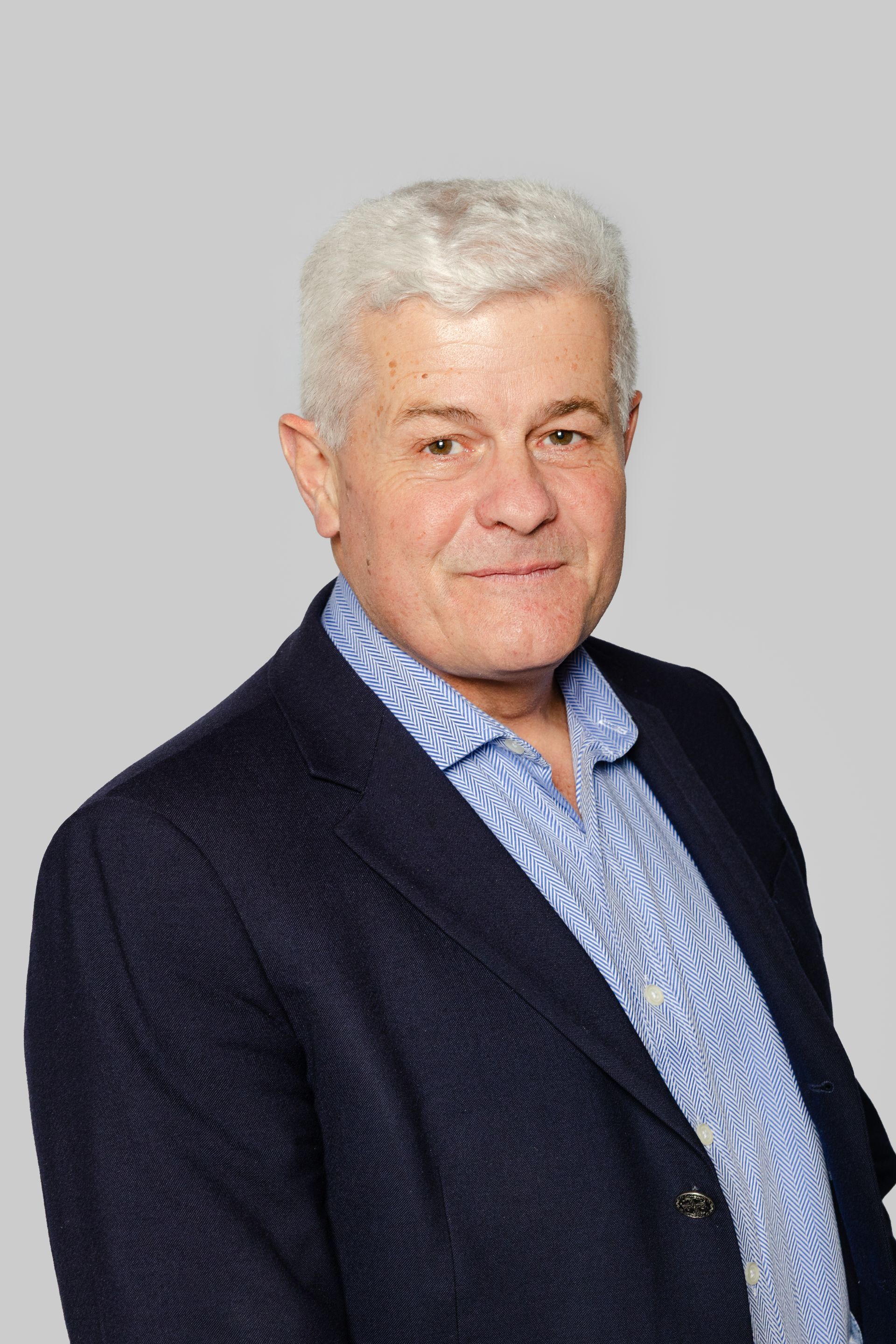 Une personne aux cheveux gris, vêtue d'une chemise bleue à motifs et d'un blazer bleu marine, pose sur un fond gris.
