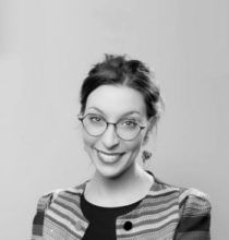 Une femme portant des lunettes et une veste à carreaux sourit sur une photo en noir et blanc.
