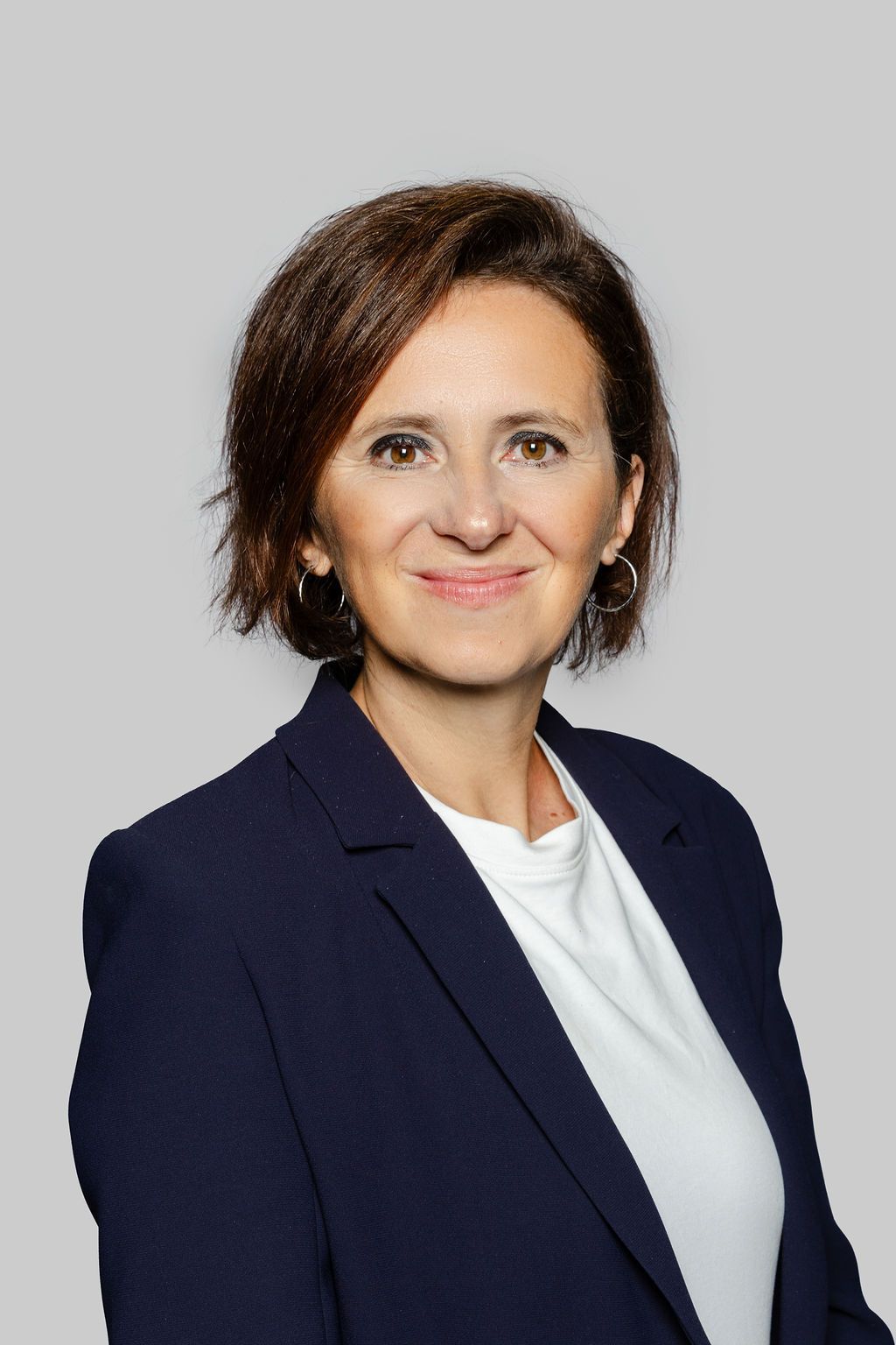Femme aux cheveux noirs et aux boucles d'oreilles créoles dans un blazer blanc et un haut noir, souriant légèrement, en intérieur.