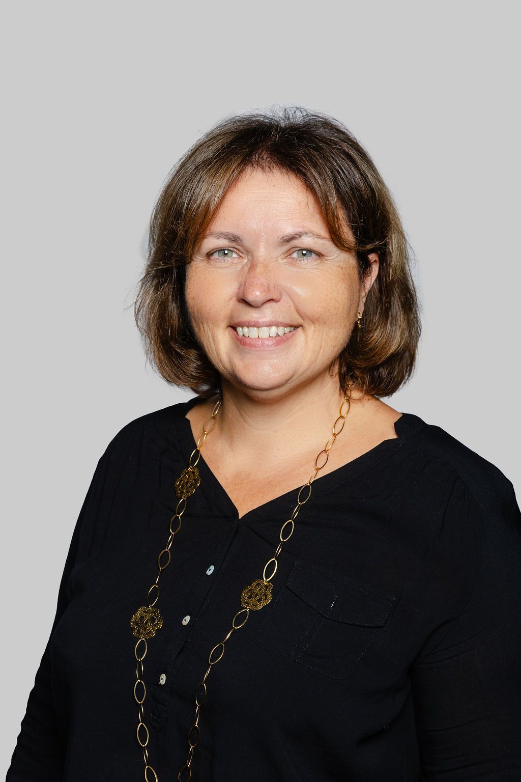 Femme souriante aux cheveux bruns mi-longs, portant un haut noir et un collier en or, sur fond gris.