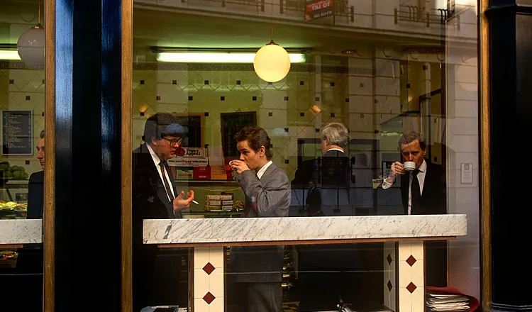 Des personnes se tiennent debout à un comptoir en marbre à l'intérieur d'un magasin, buvant dans de petites tasses, que l'on aperçoit à travers une grande vitrine.