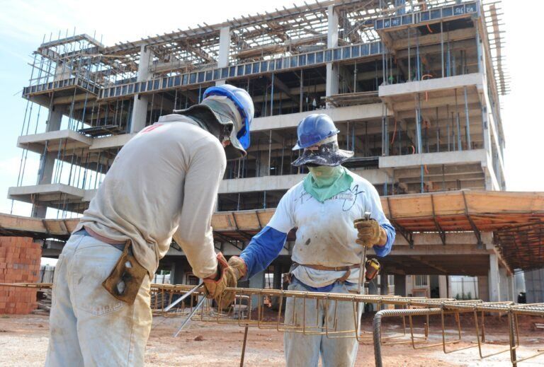 Operários da construção civil inspecionam vergalhões em um canteiro de obras, usando capacetes de segurança e luvas de trabalho.