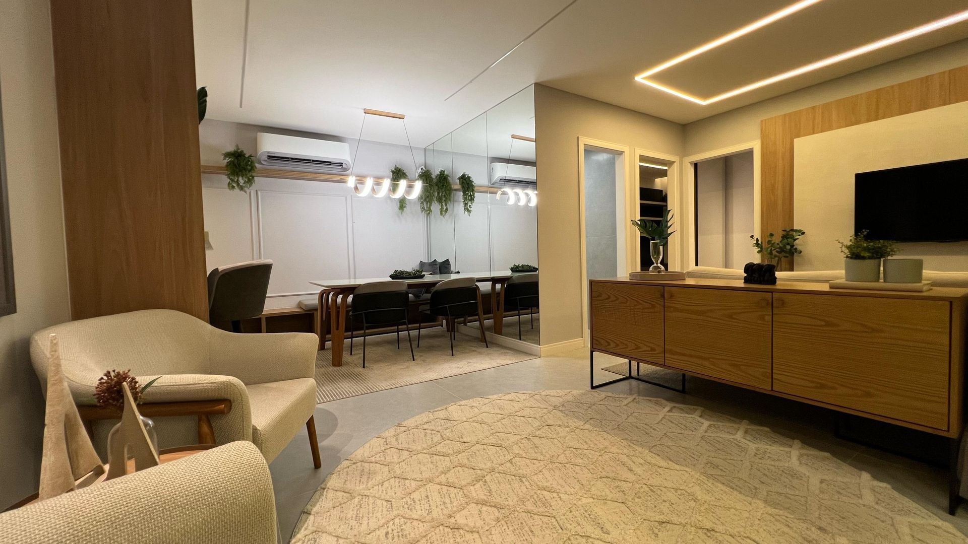 Sala de estar com poltronas creme, armários de madeira e tapete. Área de jantar visível no espelho.
