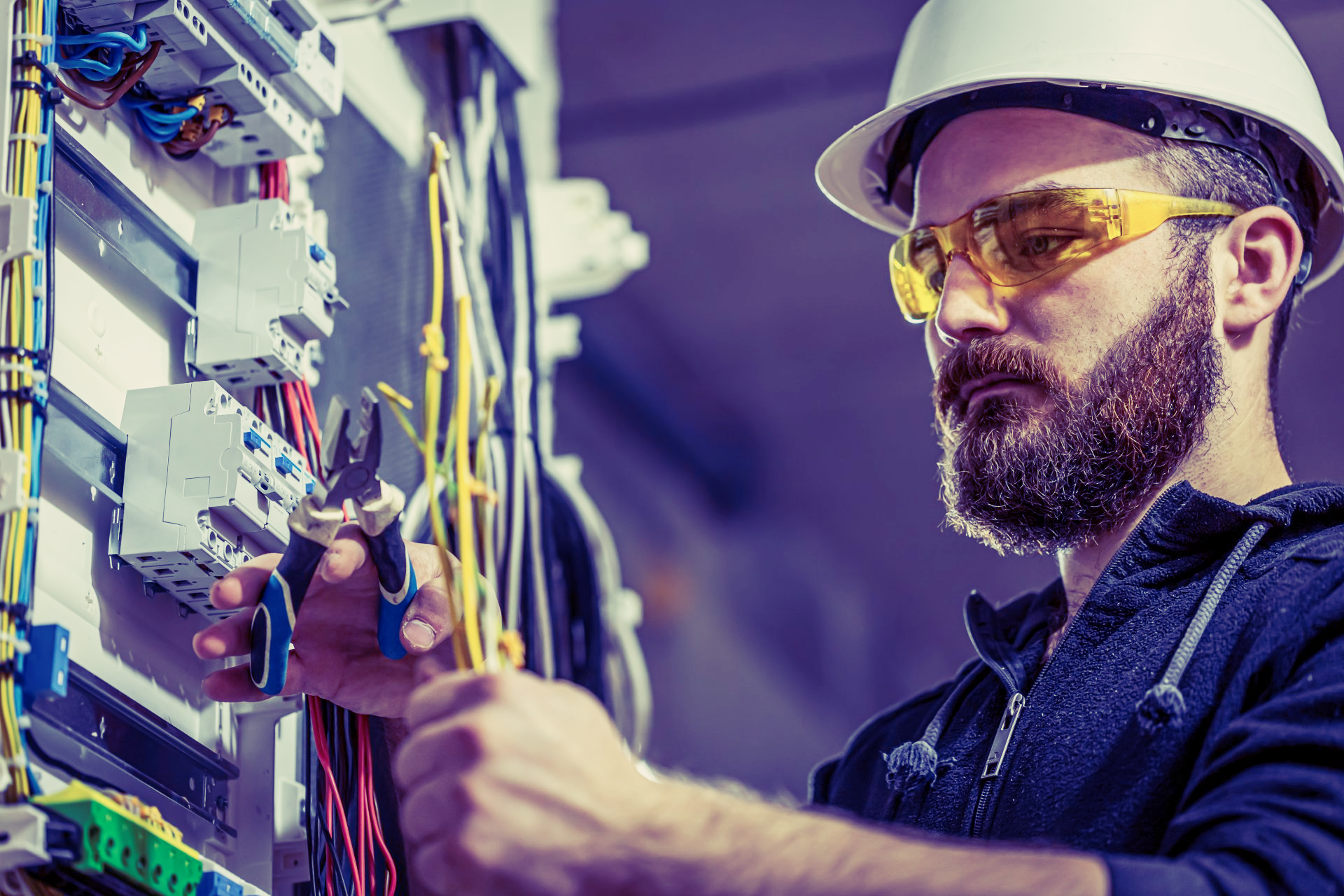 Eletricista com equipamento de segurança, trabalhando na fiação dentro de um painel elétrico.