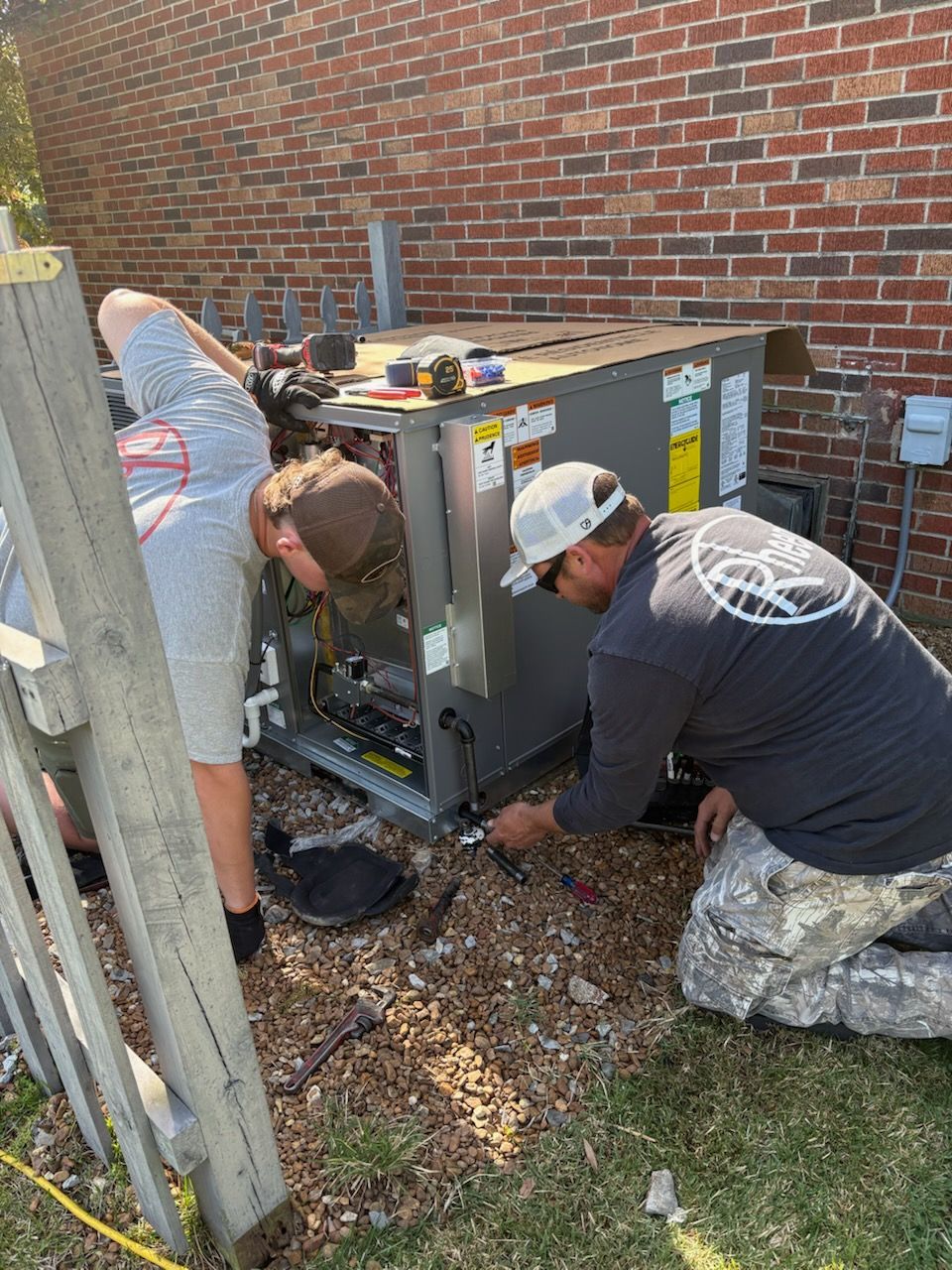 Two men are working on an air conditioner outside of a brick building.