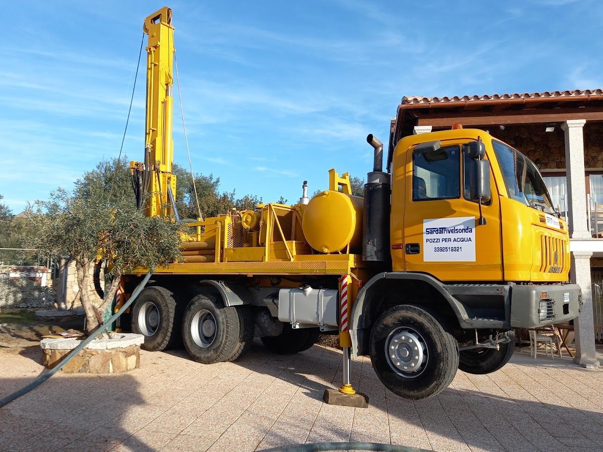 Un camion giallo con una piattaforma di perforazione parcheggiato vicino a un edificio sotto un cielo azzurro, sta perforando un pozzo Sarda Trivelsonda Group Olbia Loiri Porto San Paolo - Loiri (SS)