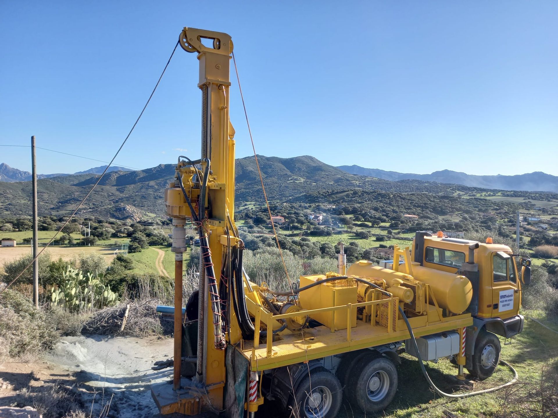 Una piattaforma di perforazione gialla su un camion in un paesaggio rurale, mentre perfora un foro sotto un cielo azzurro e terso Sarda Trivelsonda Group Olbia Loiri Porto San Paolo - Loiri (SS)