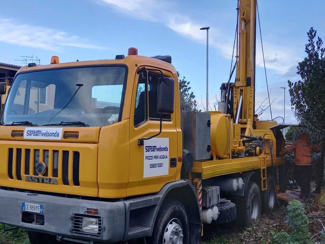Un camion di perforazione giallo parcheggiato sull'erba, accanto a un cespuglio. Un uomo lavora vicino alla piattaforma Sarda Trivelsonda Group Olbia Loiri Porto San Paolo - Loiri (SS)