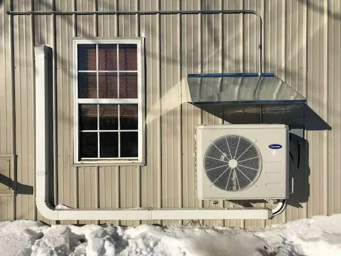 White air conditioner unit mounted on the exterior of a metal building next to a window; snow on the ground.