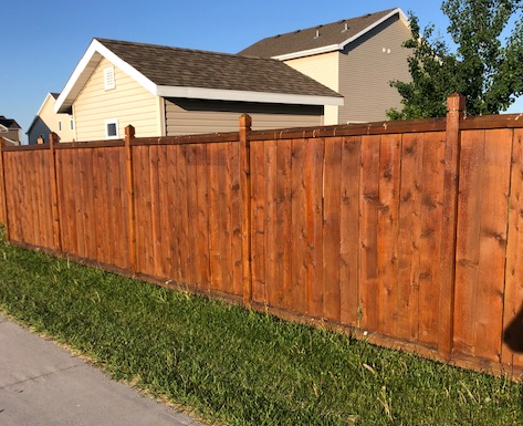 Brown wooden fence alongside a sidewalk with a beige house in the background and green grass in the foreground.