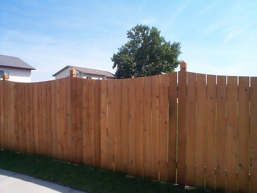 Wooden fence with dark vertical boards and decorative posts, green grass, and houses in the background.