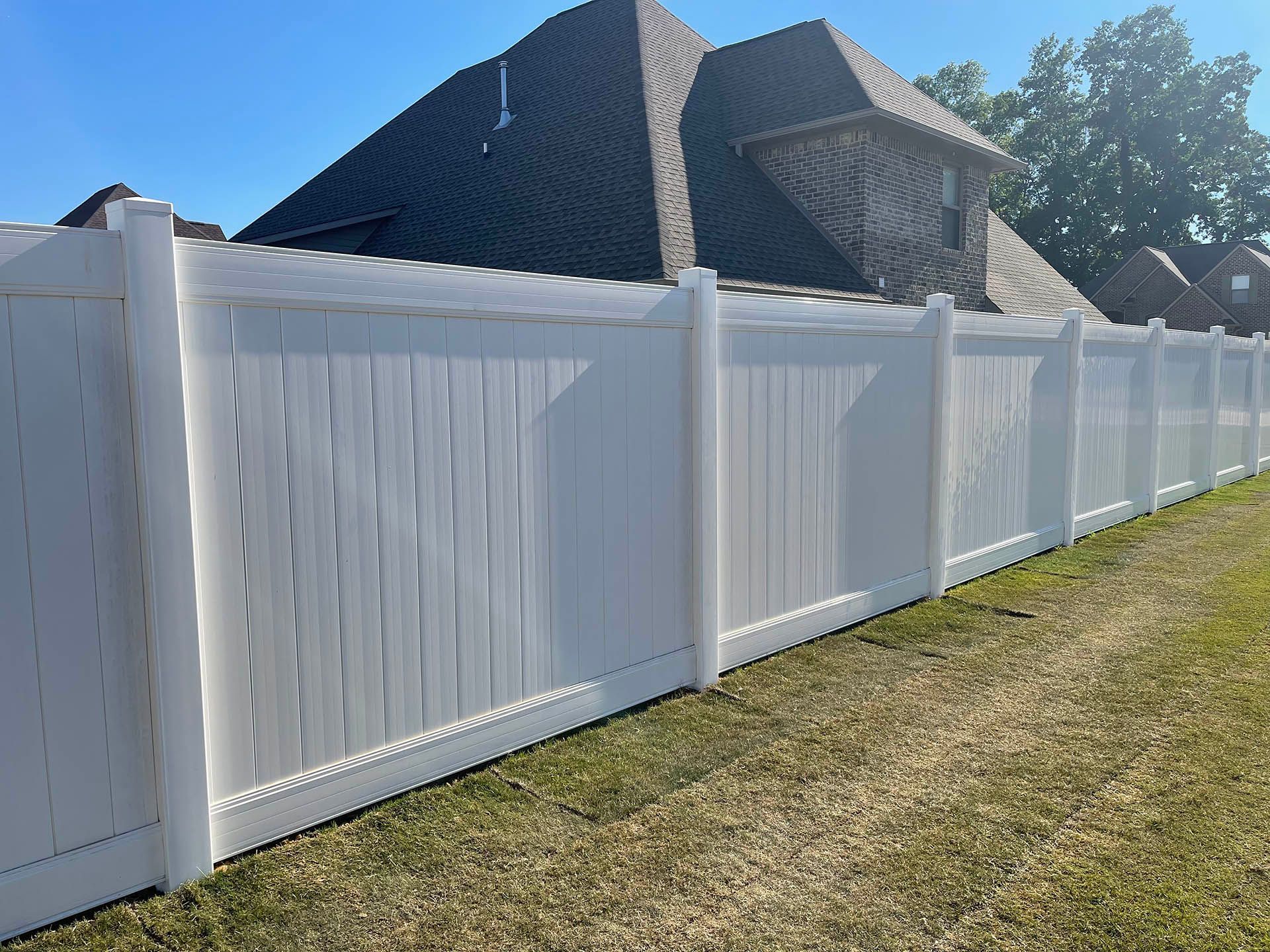 White vinyl privacy fence in front of a house with a brown shingle roof, on a sunny day.