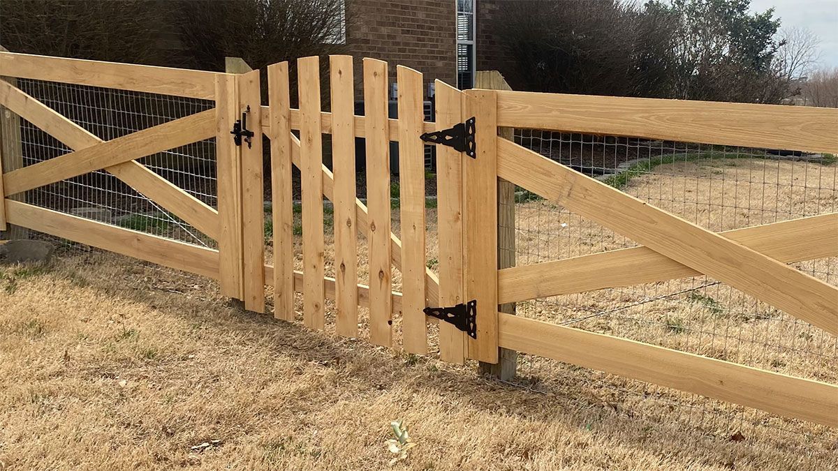 A light-colored wooden gate set within a matching split-rail fence with wire mesh, featuring black iron hinges.