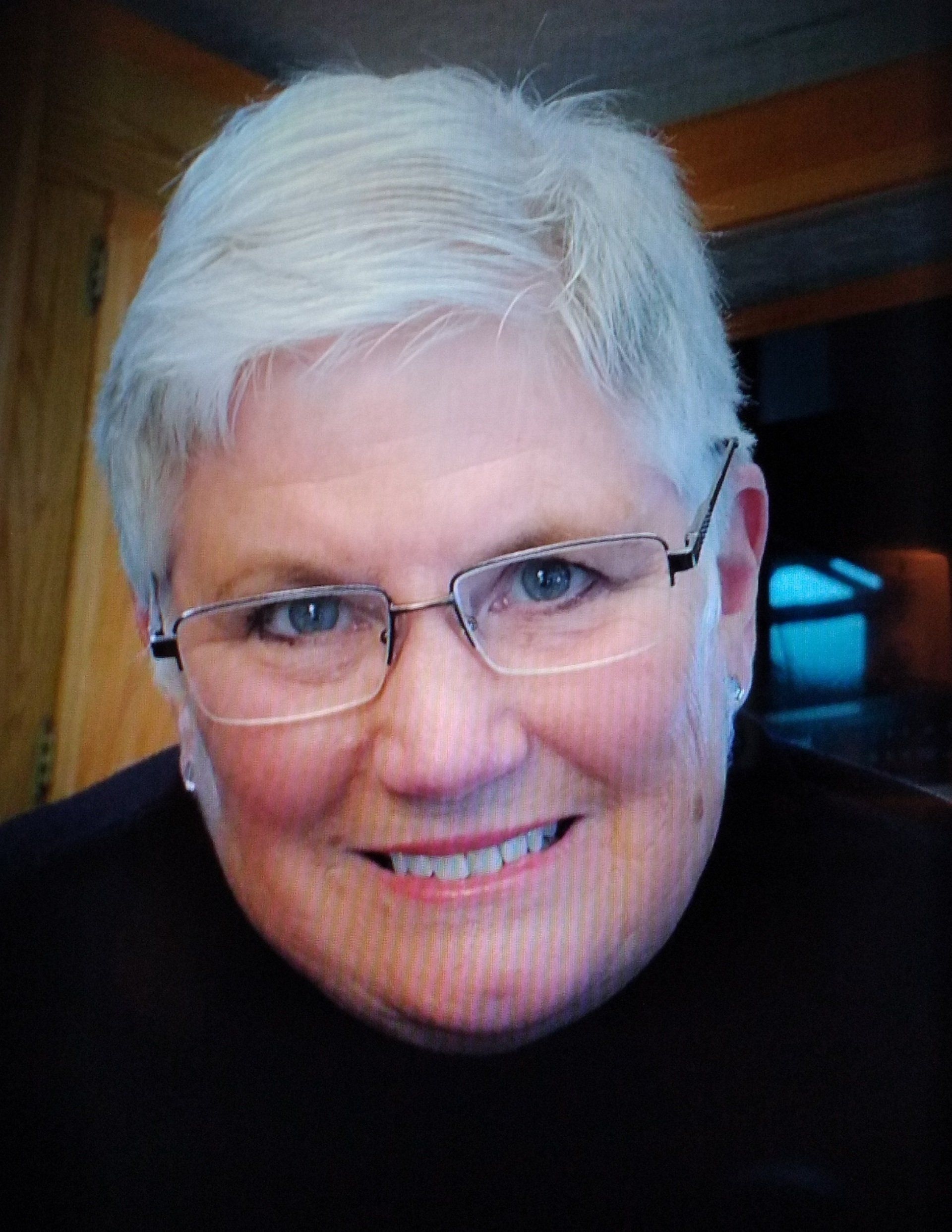 Woman with short white hair, glasses, smiling, wearing a dark top.