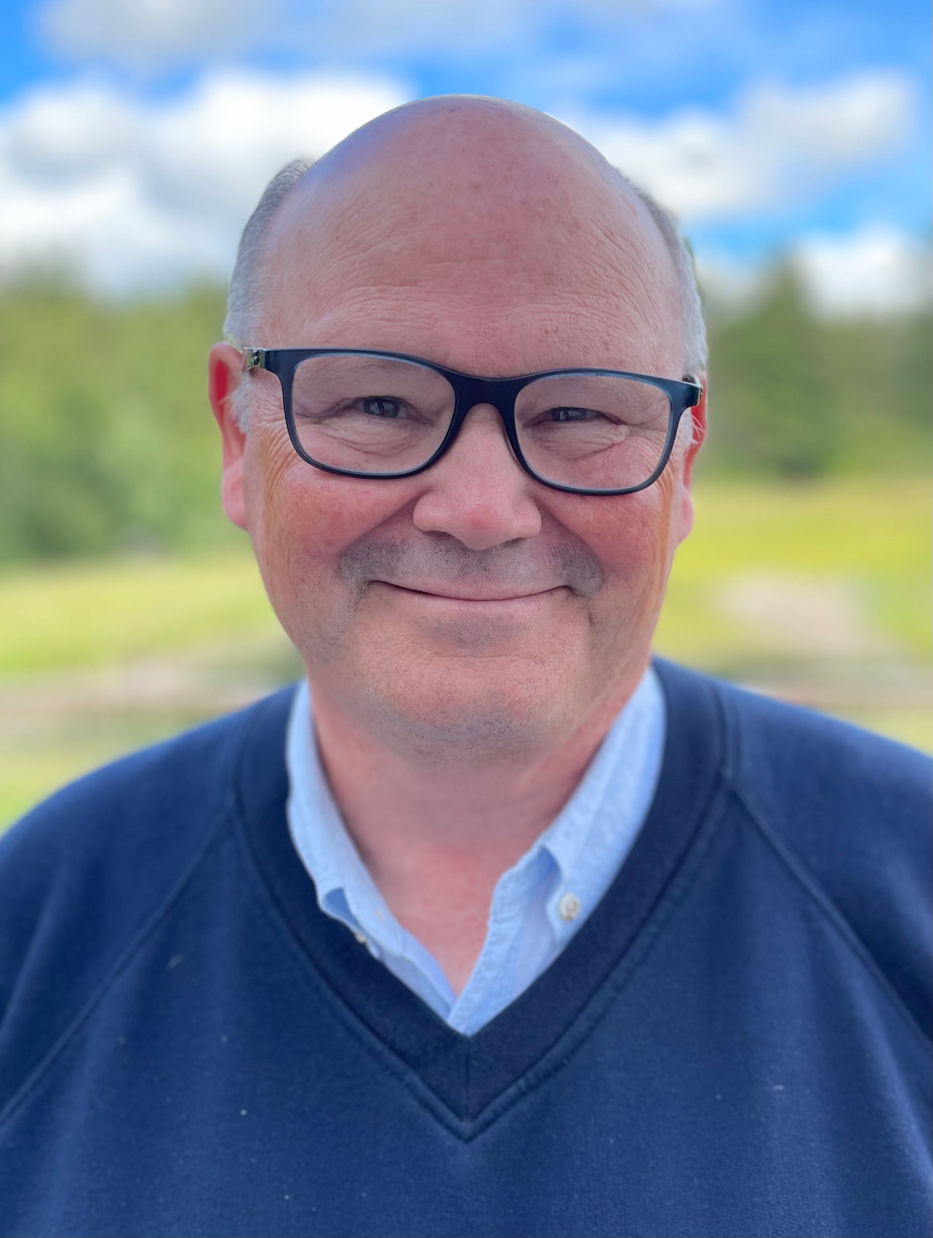 Middle-aged man with glasses, smiling, outdoors in a blue sweater and light-blue collared shirt.
