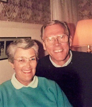 Older smiling couple indoors. Woman in teal sweater, man in glasses, dark sweater.