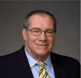 Man with glasses in suit, smiles. Dark jacket, light blue shirt, yellow tie with blue stripes.
