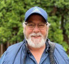 Man with glasses and white beard smiles, wearing a blue hat and jacket, outdoors.
