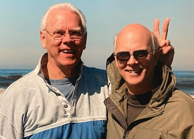 Two older men smiling at the beach; one makes a peace sign.