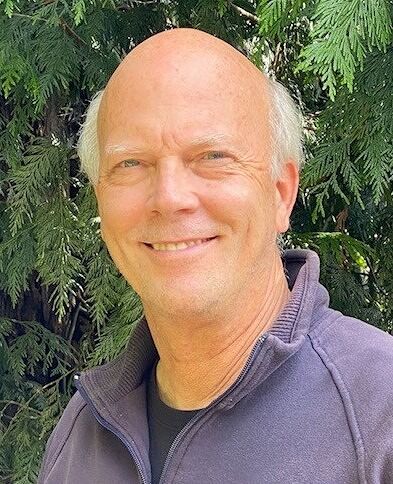 Bald man smiling, wearing purple sweater, outdoors. Green foliage in background.