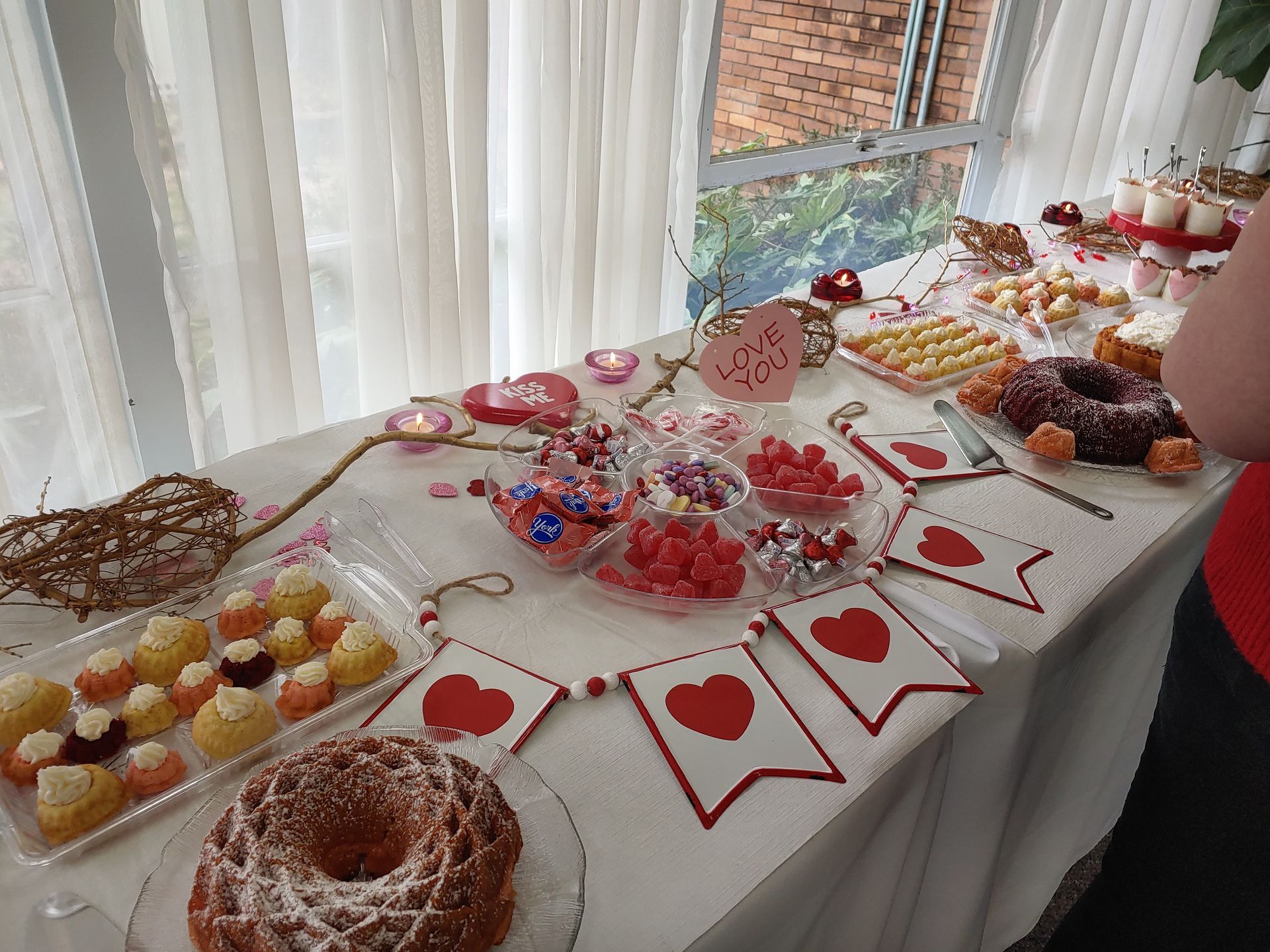 A table laden with Valentine's Day treats: cakes, cookies, and candies, decorated with heart-themed banners.