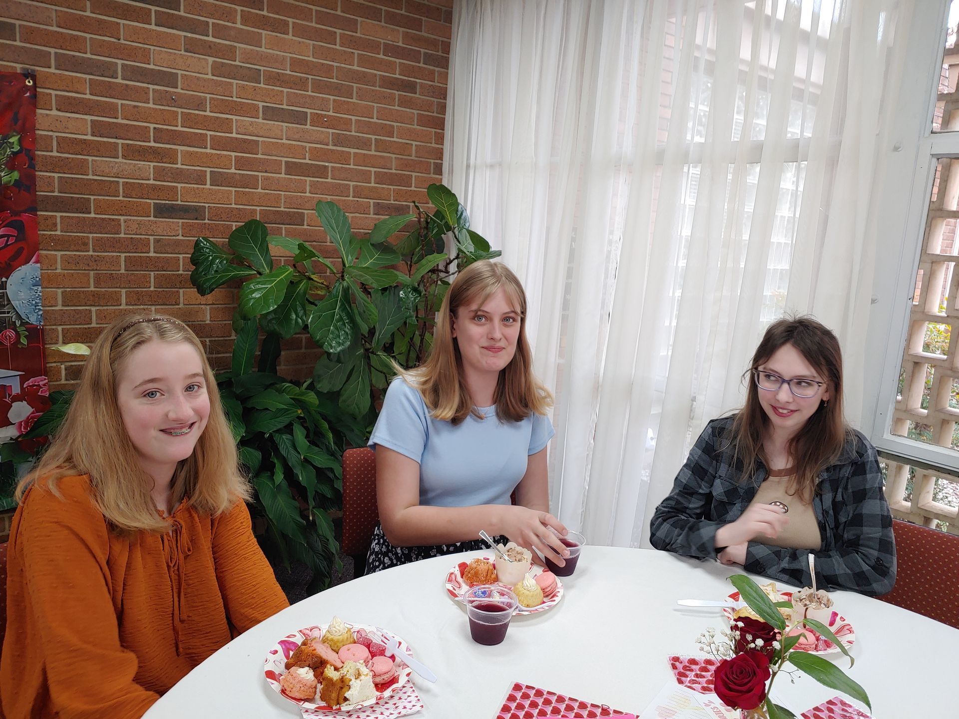 Three people seated at a round table; plates of pastries and flowers. Brick wall and window visible.