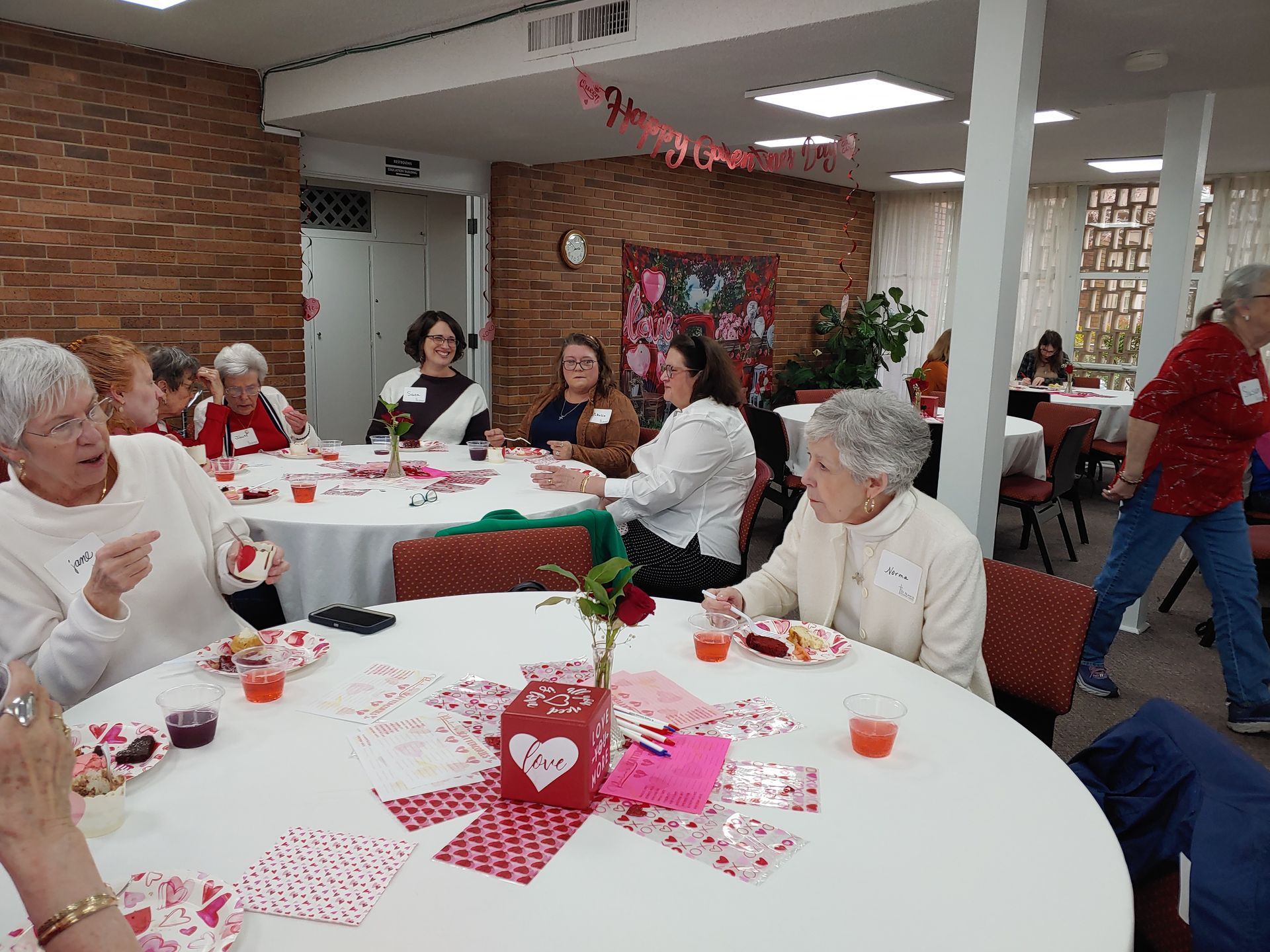 People at round tables with Valentine's Day decorations. Brick wall and white columns in the background.
