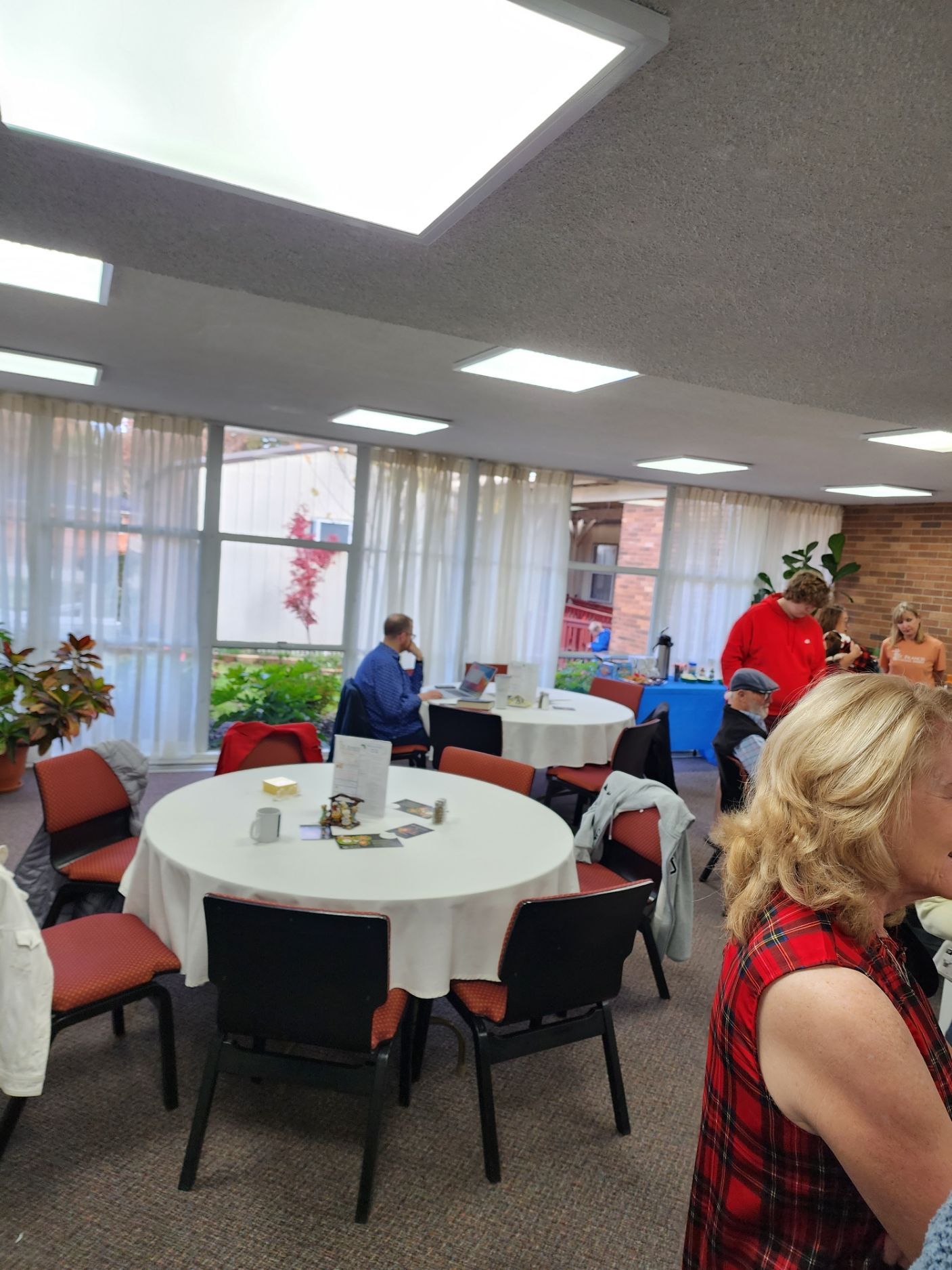 Interior room with round tables, chairs, and people; large windows and overhead lights.