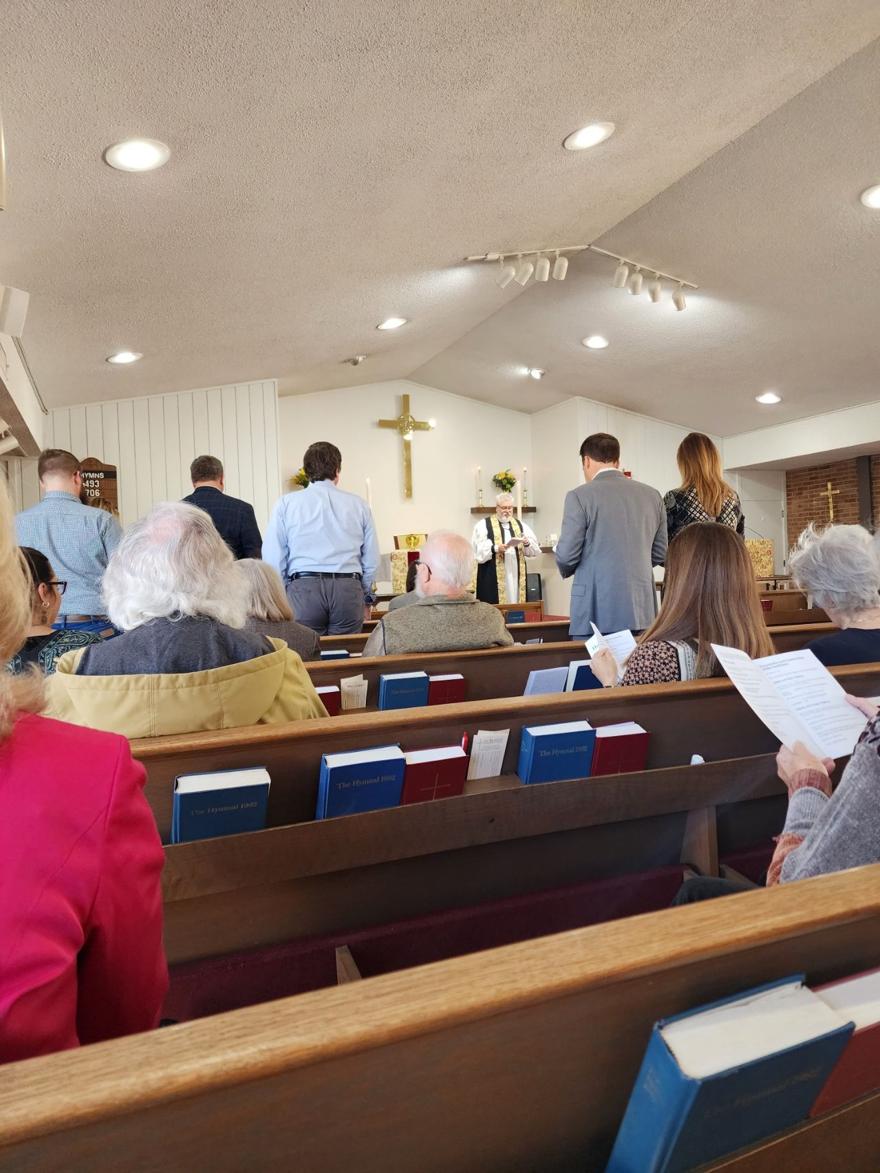People in a church service, some standing, facing a cross on the wall. Wooden pews, hymnals present.