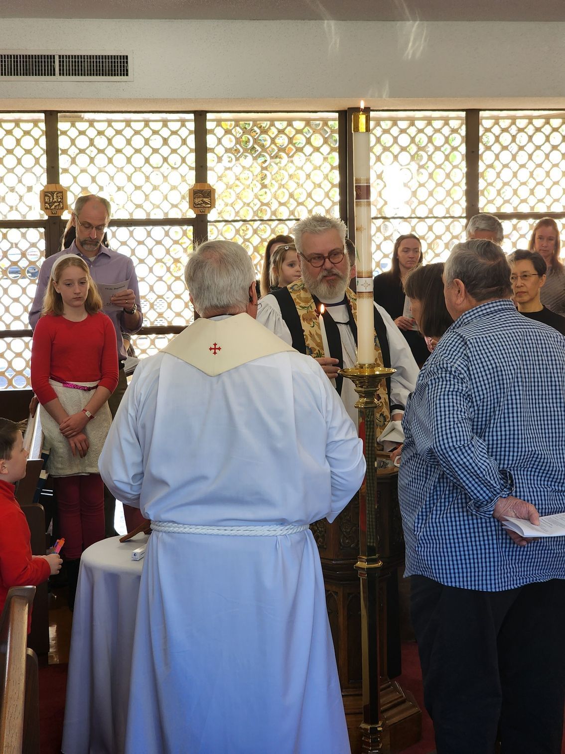 Clergy at altar with people in a church service.  A candle burns. Others stand nearby.